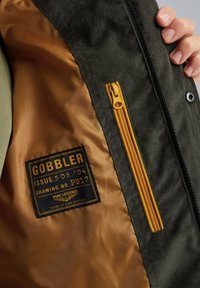 Interior de chaqueta de pana verde con forro dorado. Cuenta con un bolsillo con cremallera a la izquierda, una etiqueta negra con texto blanco y detalles de cremallera naranja.