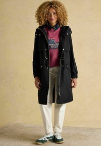 Čierny park s predným zipsom, zúženým pásom a striebornými detailmi. Nosený cez ružový sveter s kvetinovým dizajnom auta, kombinovaný s bielymi nohavicami a zelenými teniskami.