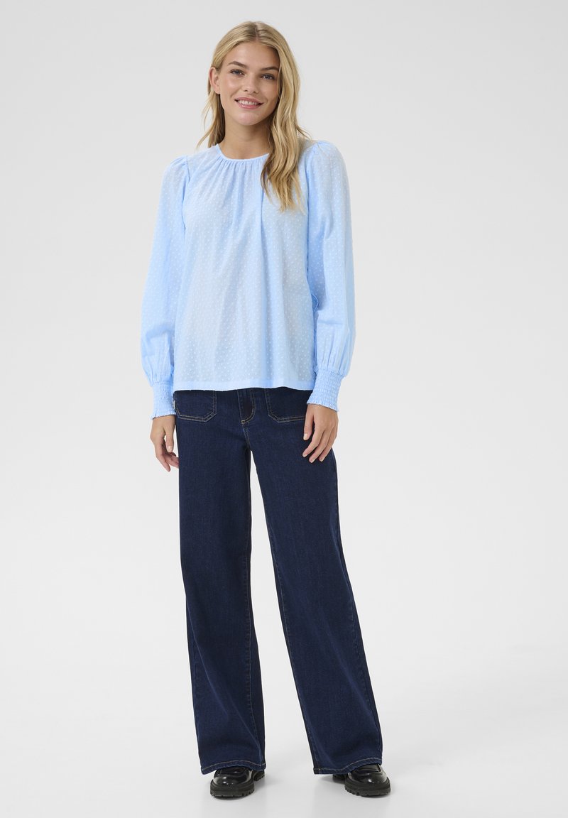 Hellbluse mit Struktur und Puffärmeln, kombiniert mit hoch sitzenden dunkelblauen Schlagjeans und schwarzem Schuhwerk.