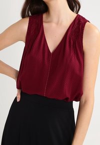 Burgundy sleeveless top with a V-neck, featuring textured fabric, embroidered detail on shoulders, and subtle front pleats.