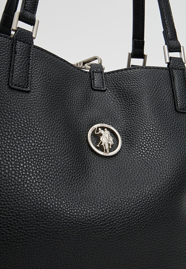 Bolso de cuero negro con una superficie texturizada, que presenta un emblema circular plateado en el centro y dos correas resistentes.
