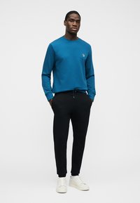 Hombre con sudadera de manga larga de color verde azulado, pantalones jogger negros y zapatillas blancas, de pie con las manos en los bolsillos frente a un fondo liso.