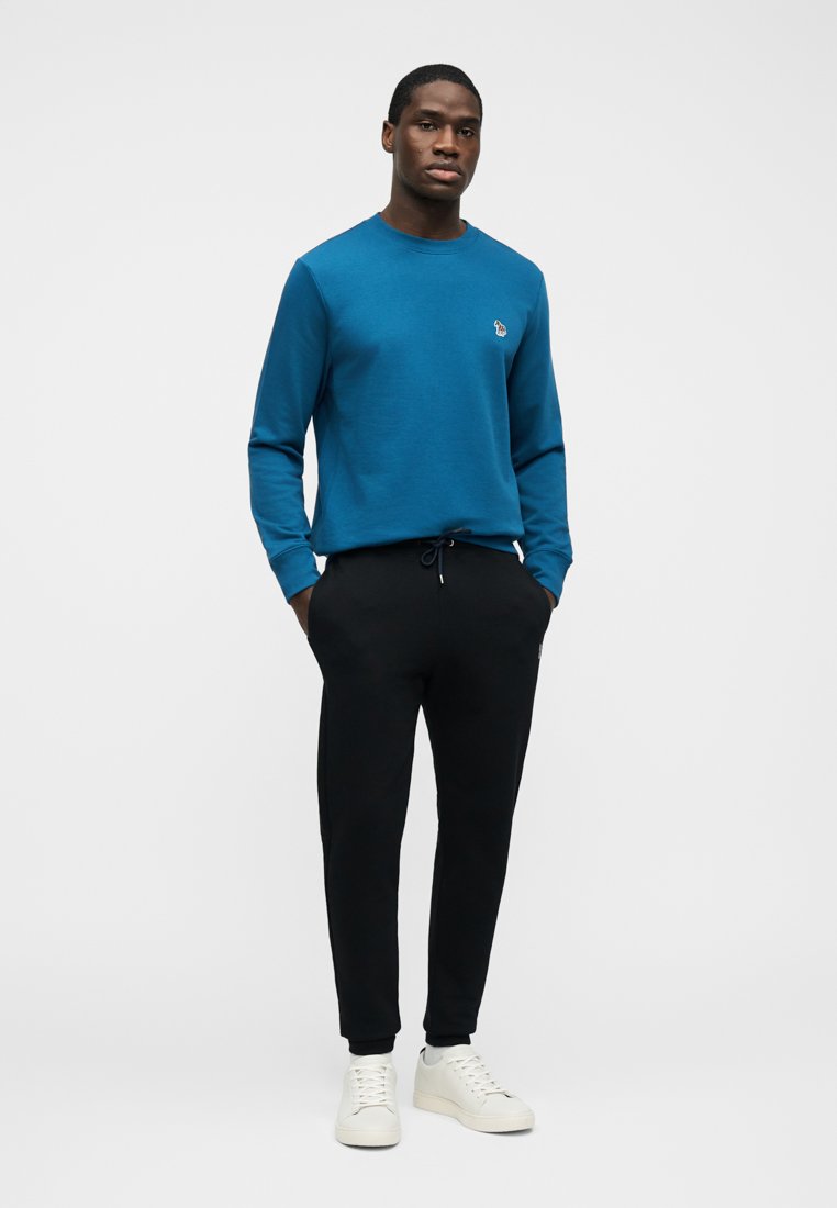 Homme portant un sweat-shirt à manches longues turquoise, un pantalon de jogging noir et des baskets blanches, debout les mains dans les poches devant un fond uni.