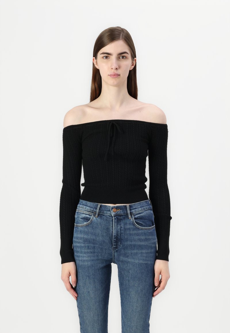 Sweater preto sem ombros com textura canelada e mangas longas, combinado com jeans de denim azul de cintura alta.