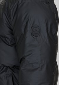 Sort pufferjakke med en glat, blank overflade, med et rundt sort logo patch på venstre ærme og quilted detaljer.