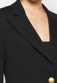 Černý blazer s ostrým klopou, strukturovanými rameny a výrazným zlatým knoflíkem. Hladká tkanina s jemným leskem a přizpůsobeným střihem.