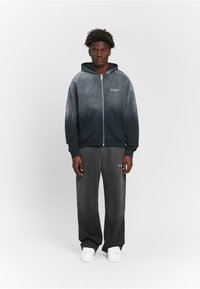 Hanorac cu fermoar de culoare gri închis, cu un finisaj gradient, având o buzunar frontal și un logo. Asortat cu pantaloni sport largi și adidași albi.