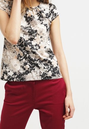 Femme portant un top à manches courtes noir et blanc à motif floral et un pantalon rouge, posant avec une main près du visage et l'autre sur le côté.