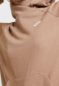 Hnědá mikina vyrobená z měkké tkaniny, s vysokým límcem, přední kapsou a bílým vyšitým logem na prsou. Hladká textura.
