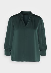Blouse vert foncé en tissu lisse, avec un col en V, des épaules froncées et des manches longues à poignets élastiques. Design à ourlet droit.
