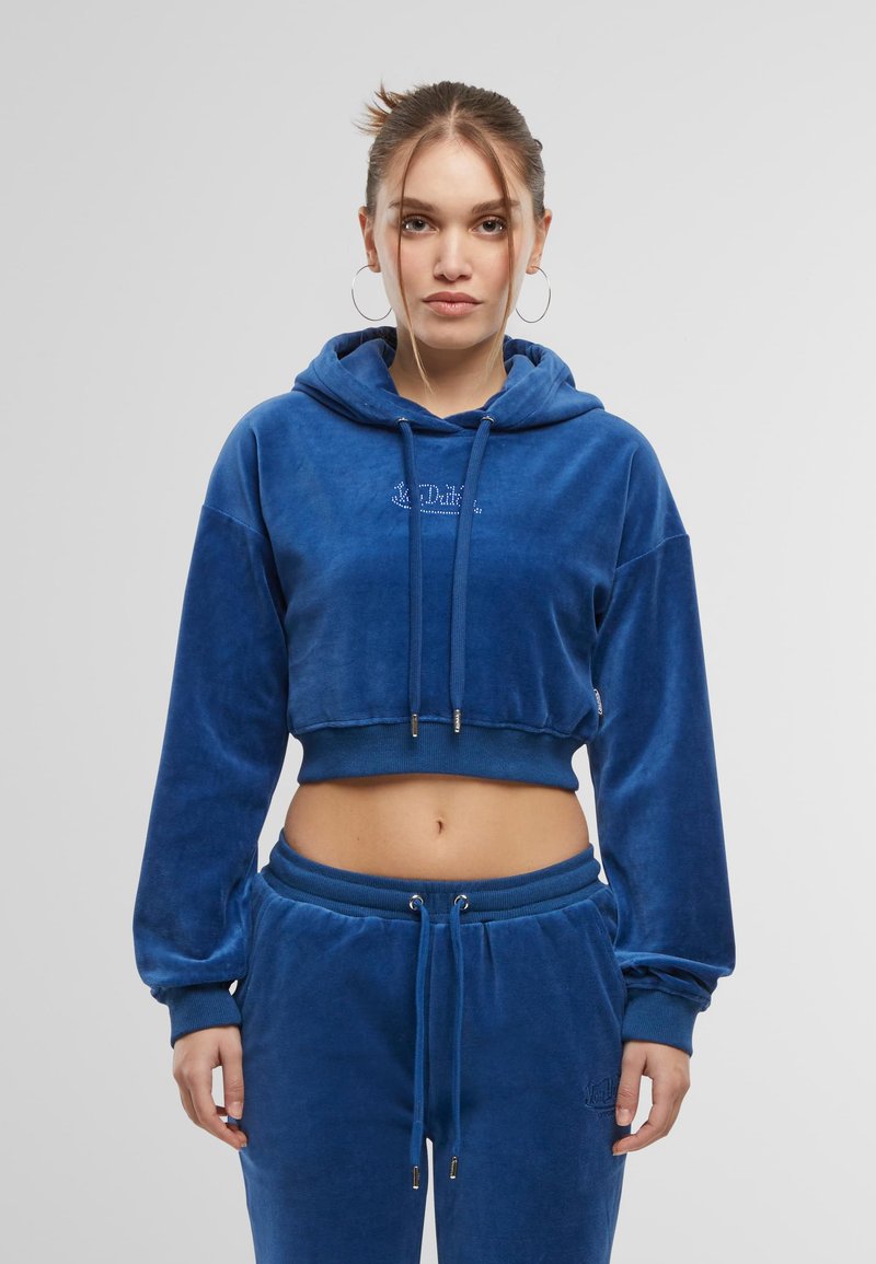 Hoodie cropped in velluto blu con cappuccio e cordino, polsini a coste e logo ricamato. Abbinato a pantaloni jogger coordinati con una texture simile.