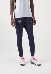 Granatowe spodnie sportowe z białym logo Nike i emblematem Anglii. Charakteryzują się zwężanym krojem, bocznymi czerwonymi akcentami oraz lekkim materiałem poliesterowym.