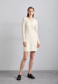 Abito a maniche lunghe di colore crema con tessuto a maglia texture, caratterizzato da un orlo liscio, scollatura rotonda e indossato con mocassini scuri.