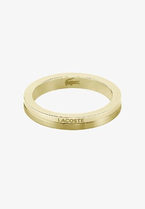 Anello in metallo color oro con superficie liscia, caratterizzato da un logo Lacoste inciso in modo sottile e da una forma rotonda. Design minimalista, senza decorazioni.
