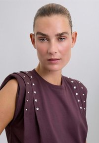 Haut bordeaux à manches courtes, avec des embellissements en clous argentés sur les épaules, fabriqué dans un tissu en mélange de coton doux.