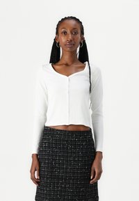 Haut cropped à manches longues blanc avec un col rond et une fermeture à boutons, associé à une jupe en tweed noire avec un motif à carreaux.