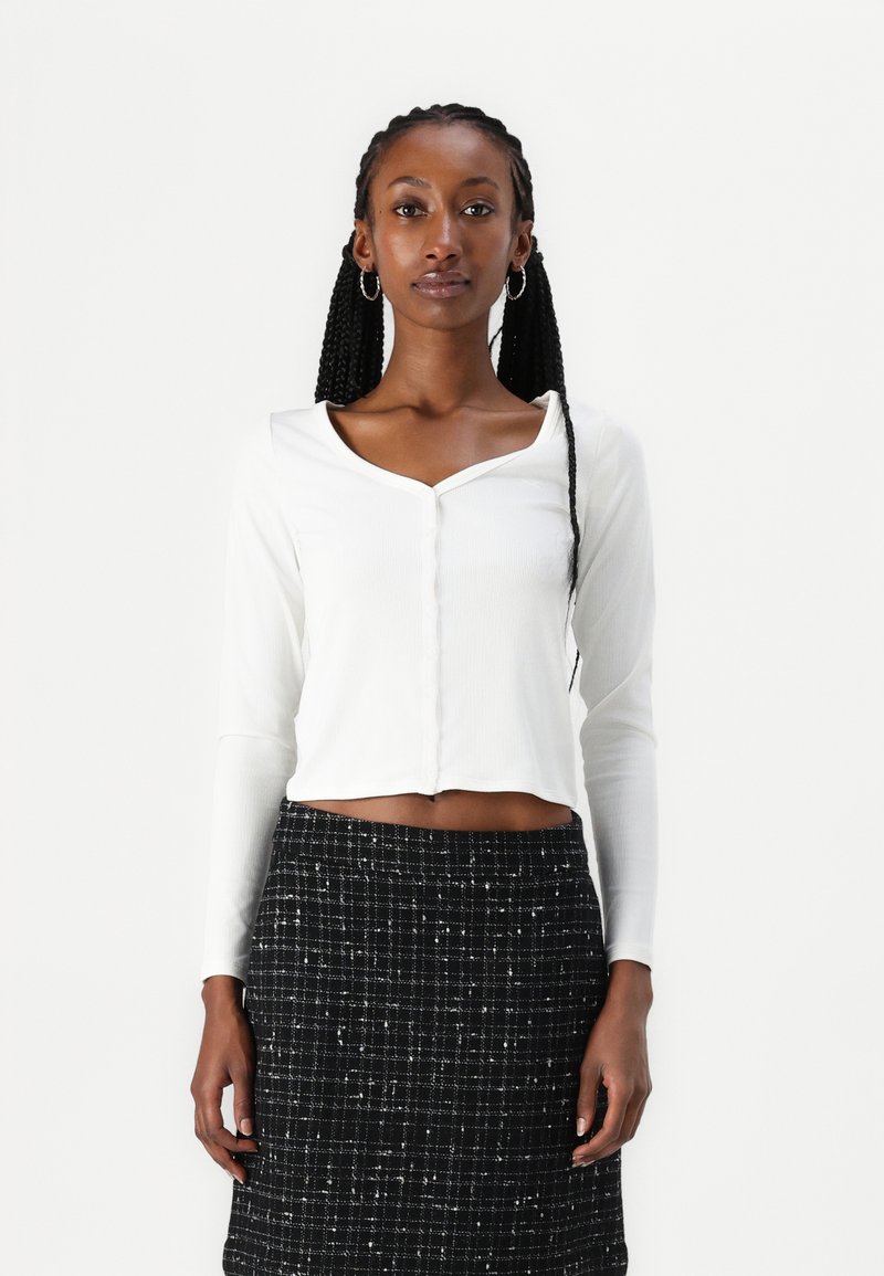 Haut cropped à manches longues blanc avec un col rond et une fermeture à boutons, associé à une jupe en tweed noire avec un motif à carreaux.