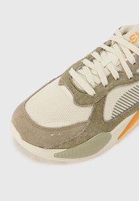 Sneaker crema e verde oliva con materiali in rete e suede, dettagli testurizzati, suola in gomma e elementi di design sovrapposti. Dettaglio di branding arancione.