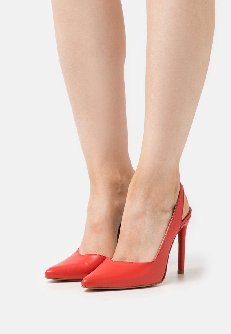 Paire d'escarpins rouges à bouts pointus avec bride arrière portés pieds nus sur un fond blanc uni.