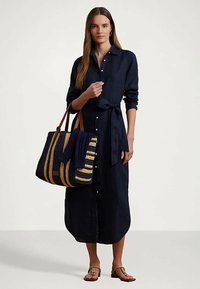 Ženska v mornarsko modri midi obleki na gumbe z vezanjem v pasu, ki nosi veliko črtasto torbo in ima na sebi rjave sandale.