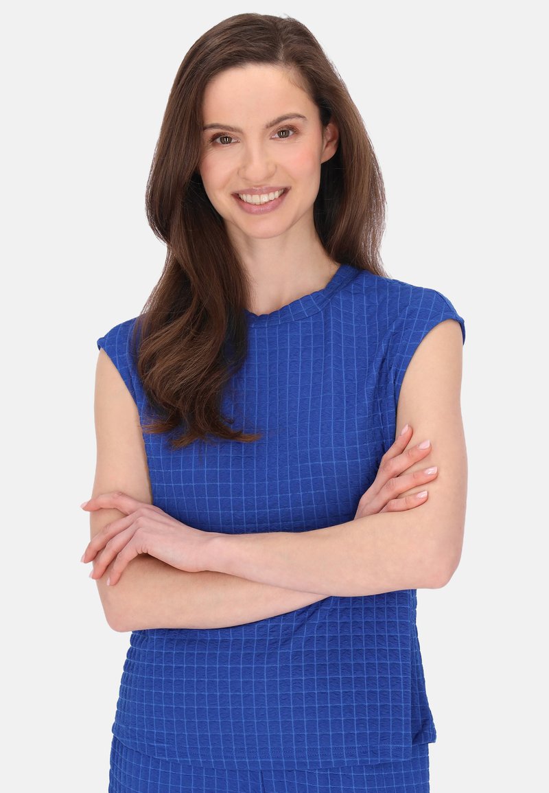 Blå texturerad topp med korta ärmar, med ett rutmönster och en rundad halsringning, bärs i en neutral miljö med armarna i kors.