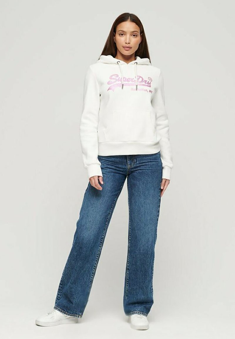 Sudadera blanca con capucha y texto gráfico en rosa, combinada con jeans de pierna ancha azul y zapatillas blancas. Diseño sencillo y casual, sin patrones.