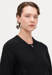 Svart texturerad jacka med rund halsringning, dragkedja framtill och sidofickor. Modellen bär stora örhängen och har en naturlig makeuplook.