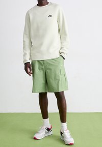 Ljusbeige sweatshirt med svart Nike-logotyp, kombinerad med gröna cargoshorts med sidofickor. Grå och rosa Nike-sneakers.