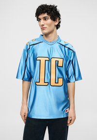 Νεαρός με σγουρά μαλλιά που φοράει μια γυαλιστερή μπλε φαρδιά μπλούζα με μεγάλα χρυσά γράμματα "IC", μαύρο παντελόνι και κολιέ με χάντρες, κοιτάζοντας αριστερά.