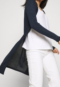 Long cardigan marine en tricot doux, avec une ouverture à l'avant et des manches longues, porté par-dessus un tee-shirt blanc et un pantalon blanc.