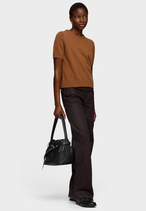 Jersey marrón de manga corta combinado con vaqueros oscuros. La modelo lleva un bolso de cuero negro y zapatos planos negros. Corte simple y relajado.