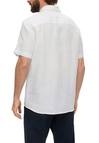 Man in een witte linnen overhemd met korte mouwen en donker gekleurde broek, gezien vanaf de achterkant tegen een witte achtergrond.
