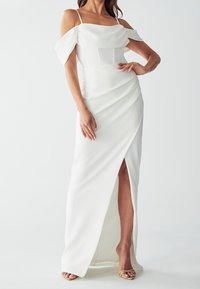 Donna in un abito da sera bianco a spalle scoperte con uno spacco alto fino alla coscia, dettagli arricciati e tacchi open-toe nudi su uno sfondo semplice.
