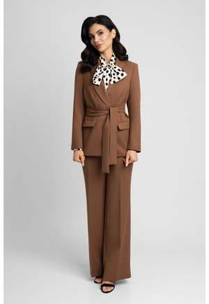 Kobieta stoi na jednolitym tle, ubrana w brązowy garnitur z paskiem, białą bluzkę w duże czarne kropki i czarne sandały na obcasie z otwartym noskiem.