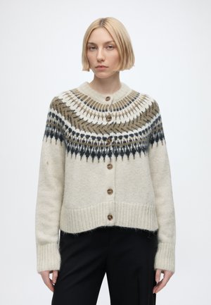 Frau mit kurzen blonden Haaren, die eine cremefarbene Strickjacke mit schwarzem und braunem gemustertem Passe und dunkle Hose trägt, steht vor einem einfarbigen Hintergrund.