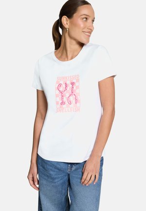 Femme portant un t-shirt blanc avec un dessin de homard rose et le texte "Sunkissed Shellfish", associée à un jean bleu, regardant sur le côté et souriant.