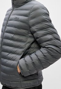 Pūšamais pelēks jaka ar augstu apkakli, horizontālām šuvēm un sānu kabatām. Uz rāvējslēdzēja atrodas logo birka. Gluda tekstūra.