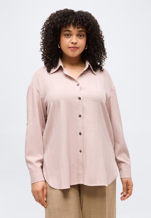 Ženska z kodrastimi lasmi, oblečena v svetlo roza srajco z gumbi in bež hlače, stoji ob nedefiniranem svetlem ozadju.