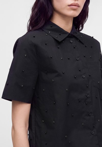 Femme portant une chemise noire à manches courtes boutonnée, décorée de petites perles noires dispersées sur le tissu et le col.