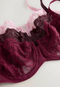 Sujetador de encaje burdeos y rosa con patrones florales, secciones transparentes y tirantes ajustables. Presenta una forma contorneada y un detalle intrincado.