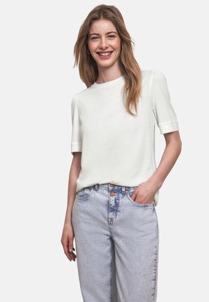 Młoda kobieta uśmiechająca się, ubrana w białą bluzkę z krótkim rękawem i jasnoniebieskie, wysoki stan jeansy, stojąca z jedną ręką w kieszeni.