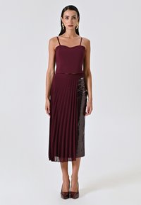 Abito due pezzi bordeaux con top aderente a spalline sottili e gonna plissettata e decorata con paillettes. Il tessuto ha una superficie liscia con riflessi scintillanti.