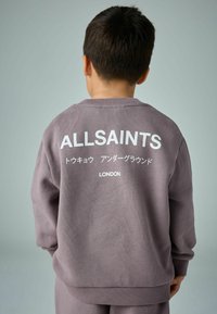 Violeta džemperis ar ribotu tekstūru, ar baltu uzrakstu "ALLSAINTS" un japāņu rakstzīmēm uz aizmugures, noapaļots kakls un nolaidām plecu šuvēm.