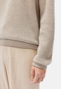 Beige gestrickter Pullover mit einem strukturierten, rautenartigen Muster, mit gerippten Bündchen und Saum. Getragen mit hellbeigen Hosen.