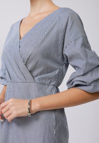 Dryžuota suknelė su giliu V formos iškirpimu ir pūstomis rankovėmis. Mėlynos ir baltos spalvos sudaro vertikalias juosteles, su apgintu liemeniu ir minkšta tekstūra.