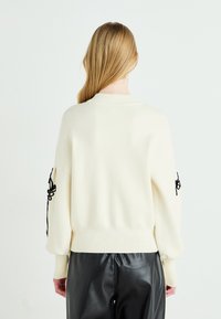 Kremfarget genser med svart broderi på ermene, med rund hals og ribbede mansjetter, parret med svarte skinnbukser.
