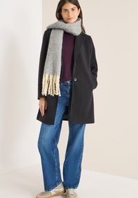 Czarny płaszcz z dwoma guzikami, purpurowy top, szerokie niebieskie dżinsy i jasnoszary szalik z frędzlami. Model stoi na jasnym tle.