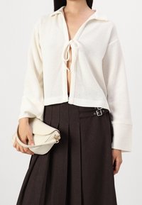 Bílý pletený kardigan s límečkem a předním zavíráním na mašličku, zkombinovaný s hnědou plisovanou sukní. Modelka drží malou bílou kabelku.