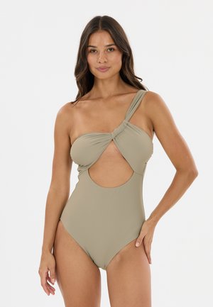 Kvinde iført beige ét-skulder badedragt med en udskæring foran, stående mod en hvid baggrund.