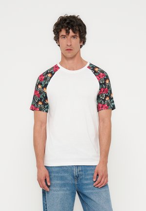 Mężczyzna z krótkimi kręconymi ciemnymi włosami, ubrany w biały t-shirt z krótkimi czarnymi rękawami w kwiaty oraz niebieskie dżinsy, stojący na jednolitym tle.